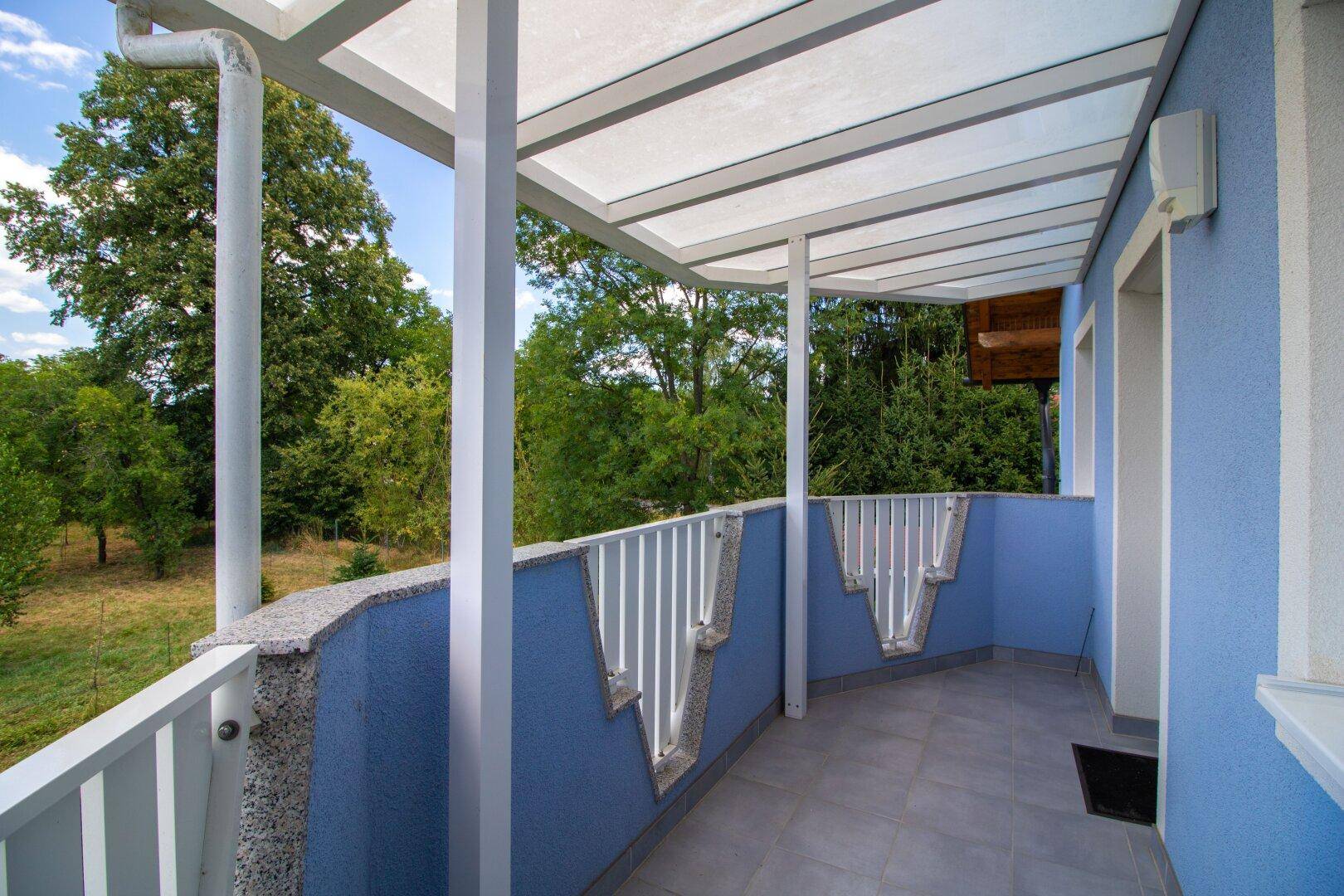 Überdachter Balkon mit blauer Fassade und Blick ins Grüne, ideal zum Entspannen im Freien.