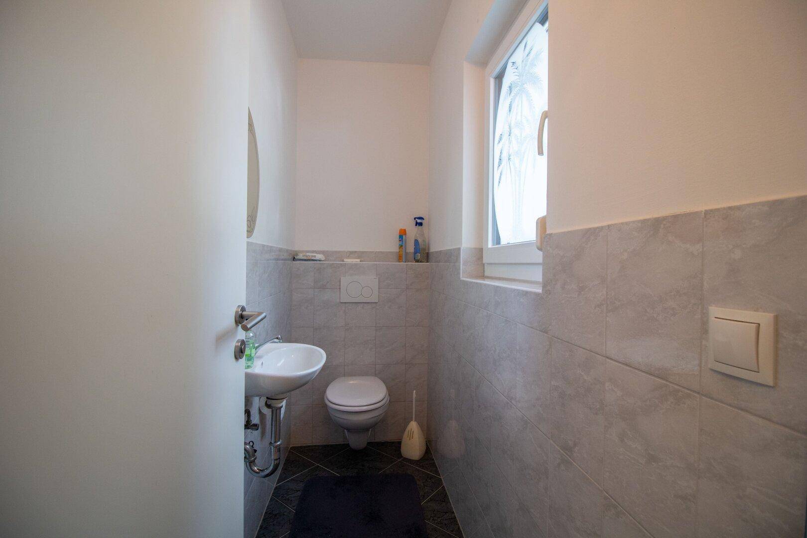 Kleines Gäste-WC mit Fenster, Wand-WC und modern gefliesten Wänden in Grau.