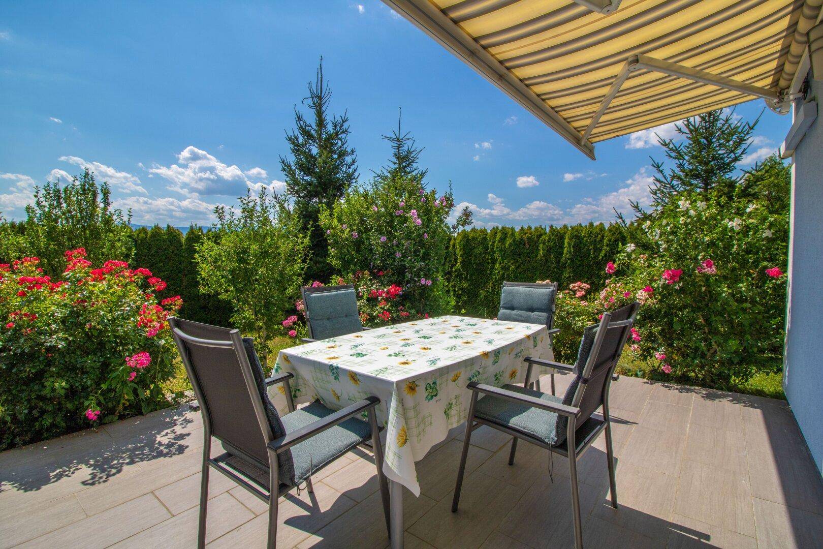 Sonniger Terrassenbereich mit Gartenmöbeln, umgeben von grünen Hecken und Blumen.