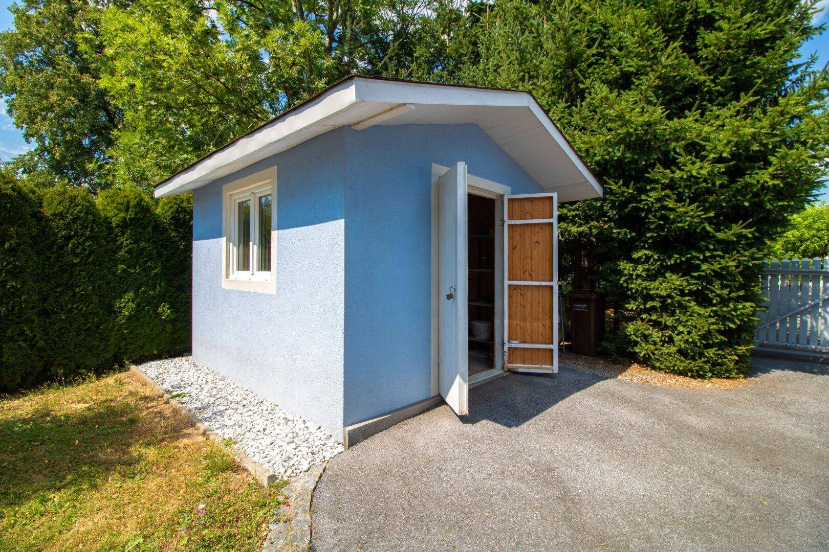 Kleines Gartenhaus mit blauer Fassade und geöffneter Holztür.