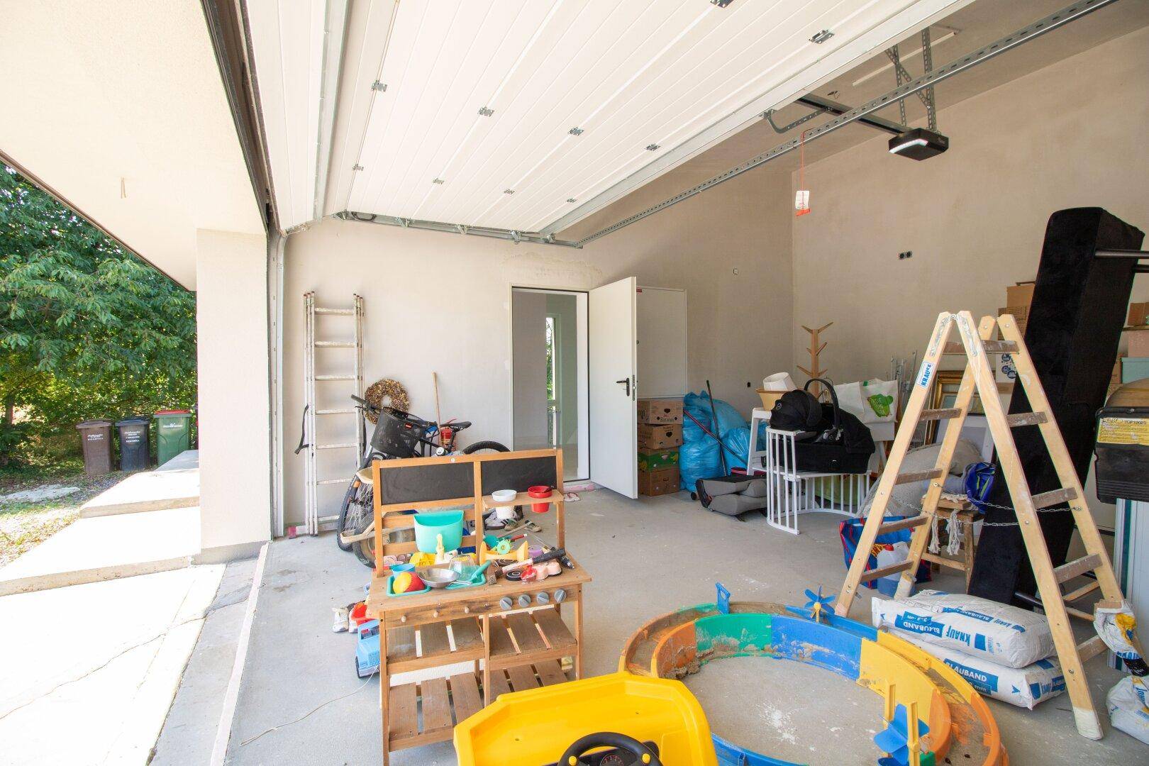 Innenansicht einer Garage mit offenem Tor, gefüllt mit Spielzeug, Leitern und Baumaterialien.