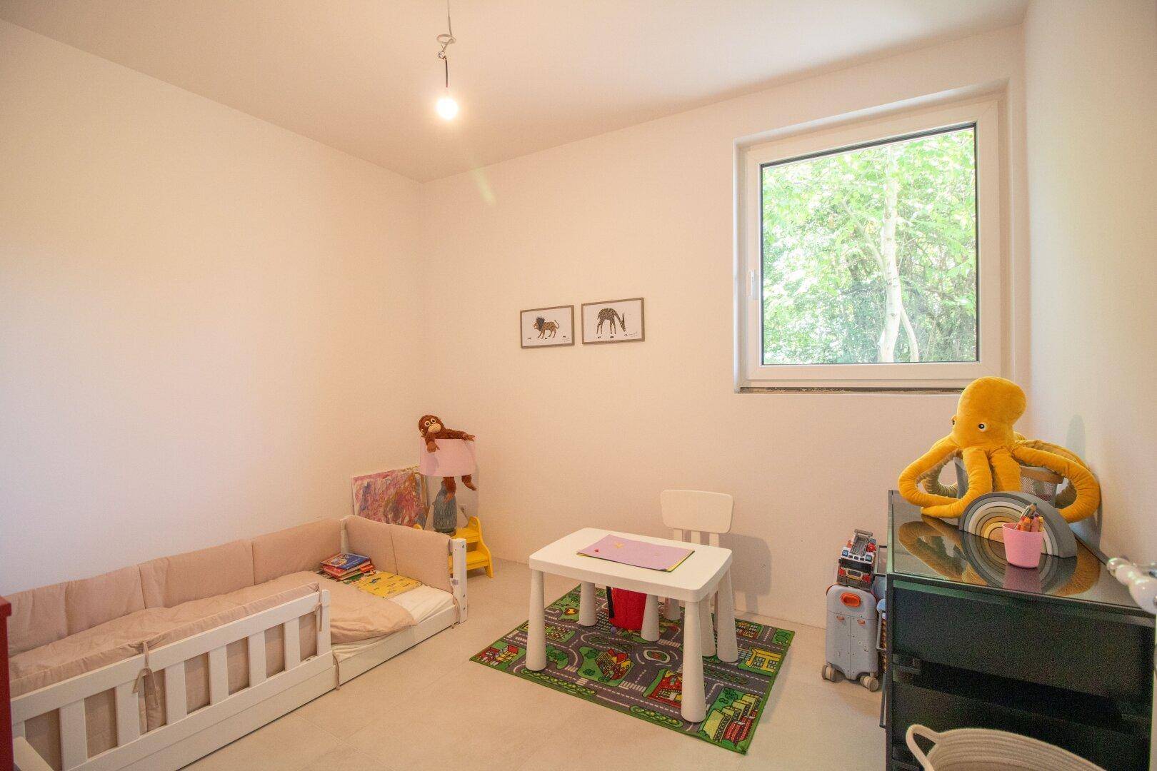 Helles Kinderzimmer mit einem kleinen Bett, Spielzeug und einem Fenster mit Blick ins Grüne.