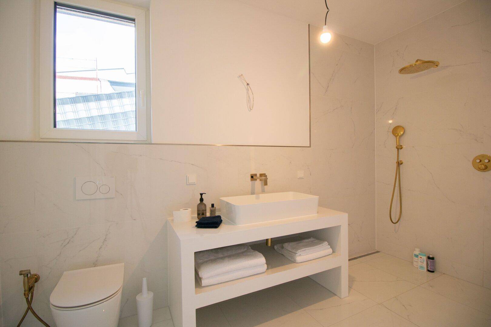 Modernes Badezimmer mit weißen Marmorfliesen, wandhängendem WC und goldfarbenen Armaturen.
