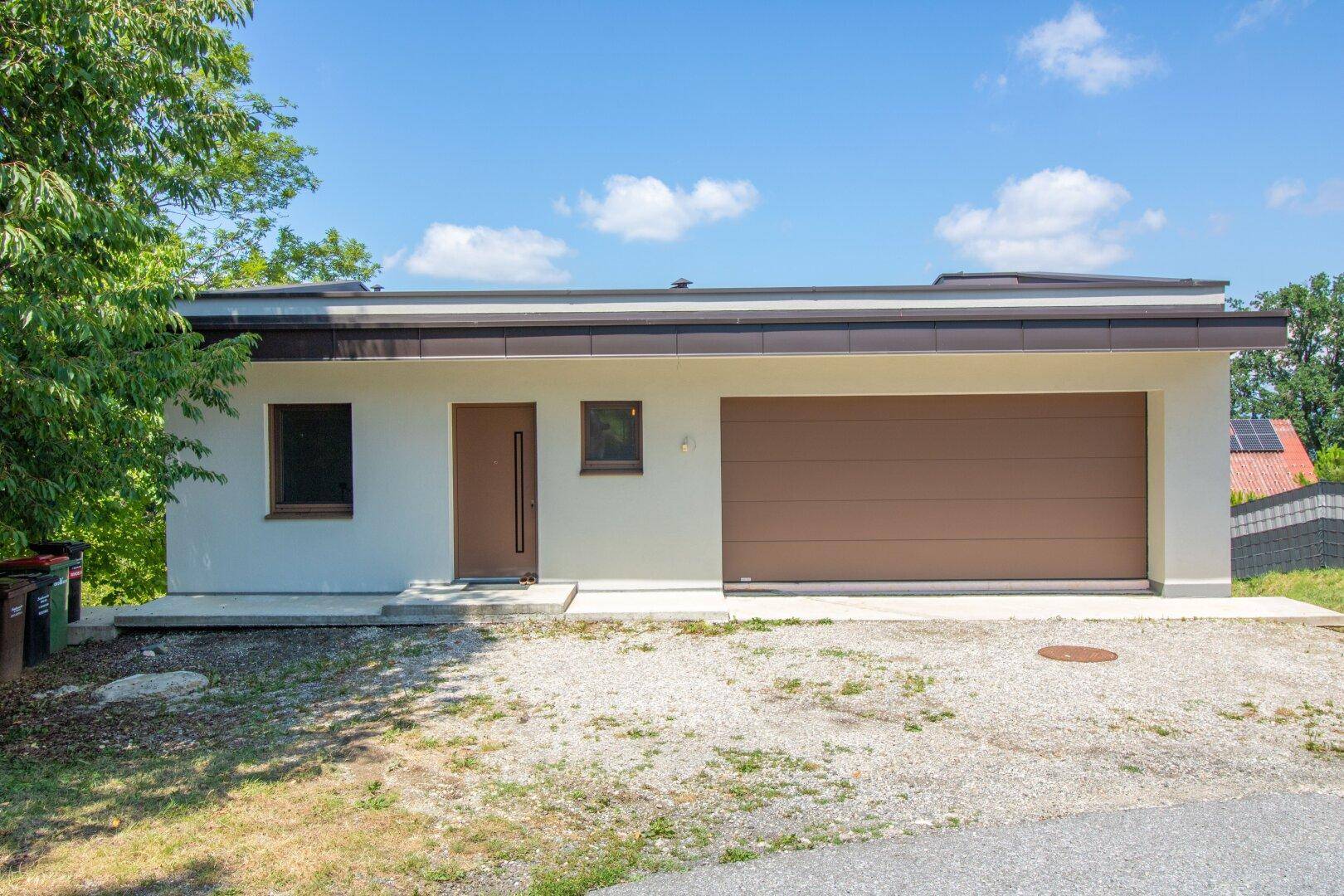 Moderne Hausfassade mit Garage, Eingangstür und flachem Dach in ländlicher Umgebung.