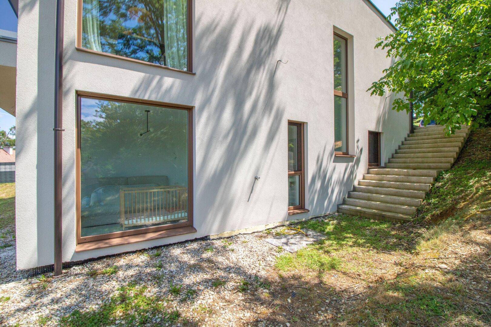 Moderne Hausfassade mit großen Fenstern und Außentreppe, umgeben von Grün.