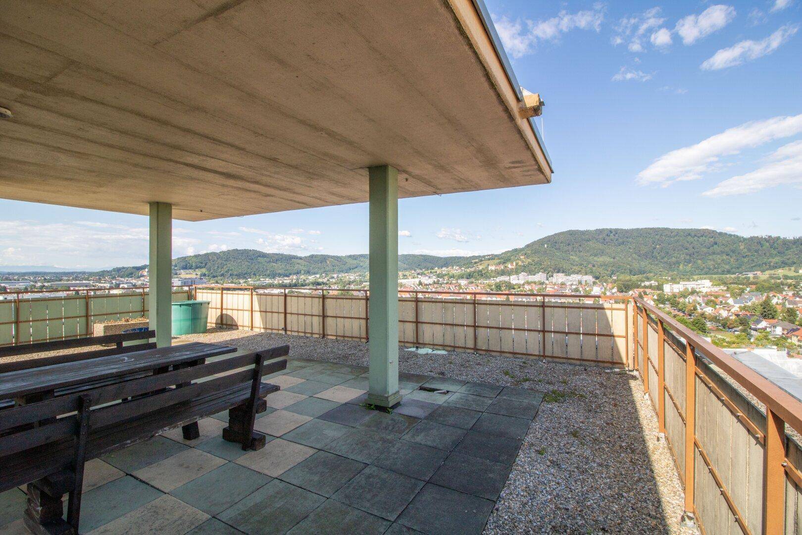 Gemeinschaftliche Dachterrasse mit Holzbänken und weitem Blick über die Stadt und die Berge.