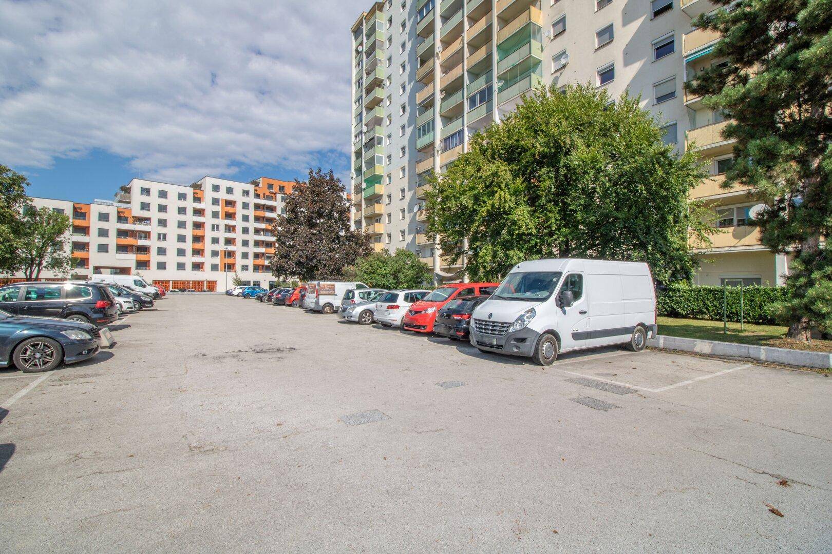 Großer Außenparkplatz vor einem Wohngebäude mit vielen geparkten Fahrzeugen und Bäumen.