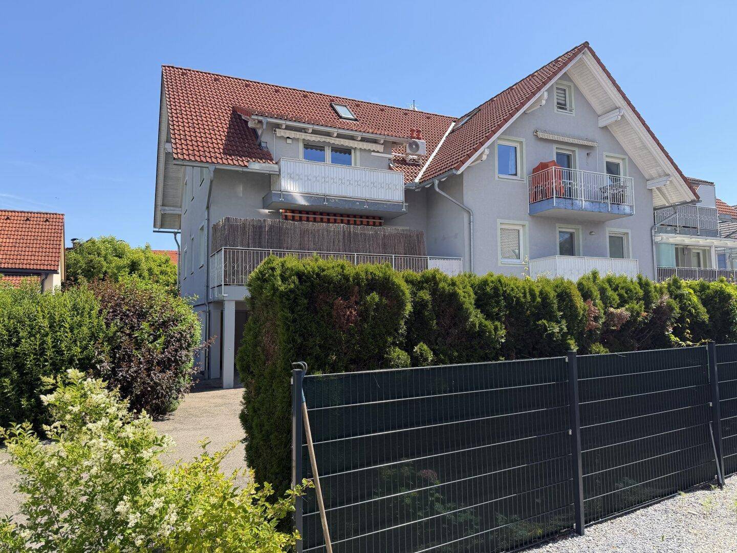Mehrfamilienhaus mit grauer Fassade, roten Ziegeldächern und mehreren Balkonen, umgeben von grünen Hecken.