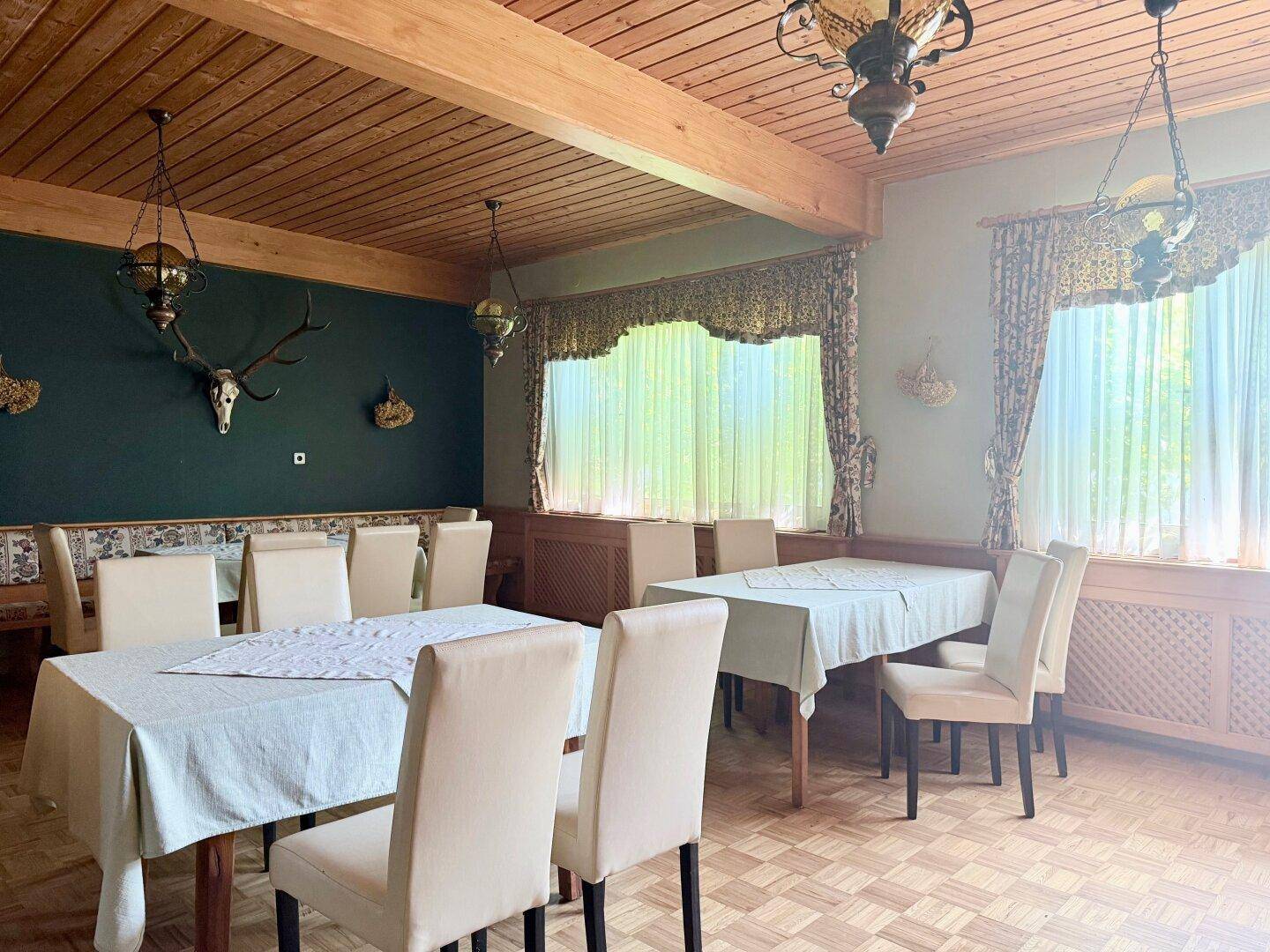 Traditioneller Speisesaal mit Holzbalken, Hirschgeweih und großen Fenstern mit Vorhängen.