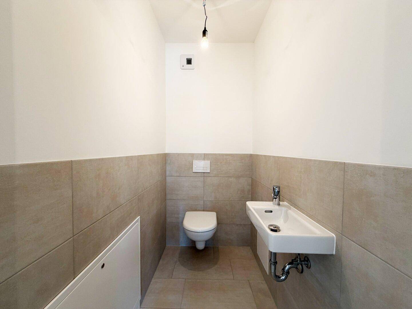 Modernes Gäste-WC mit Wand-WC, kleinem Waschbecken und hellen Fliesen an den Wänden.