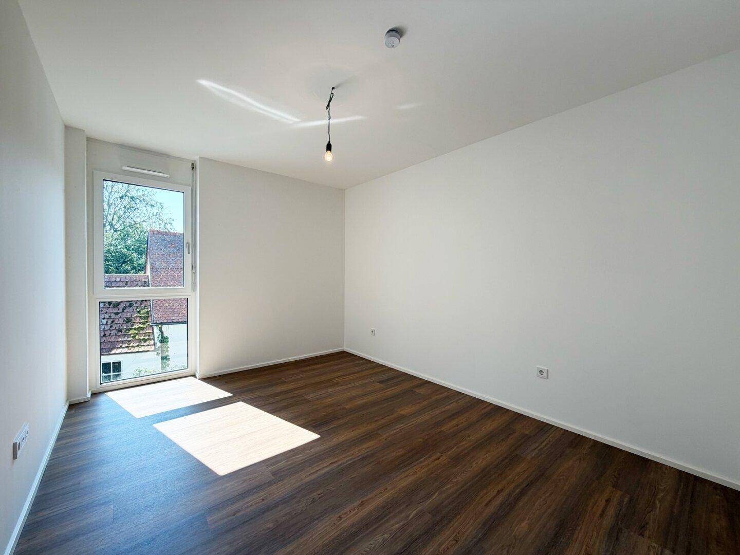 Helles Schlafzimmer mit Laminatboden und großem Fenster, das viel Tageslicht hereinlässt.