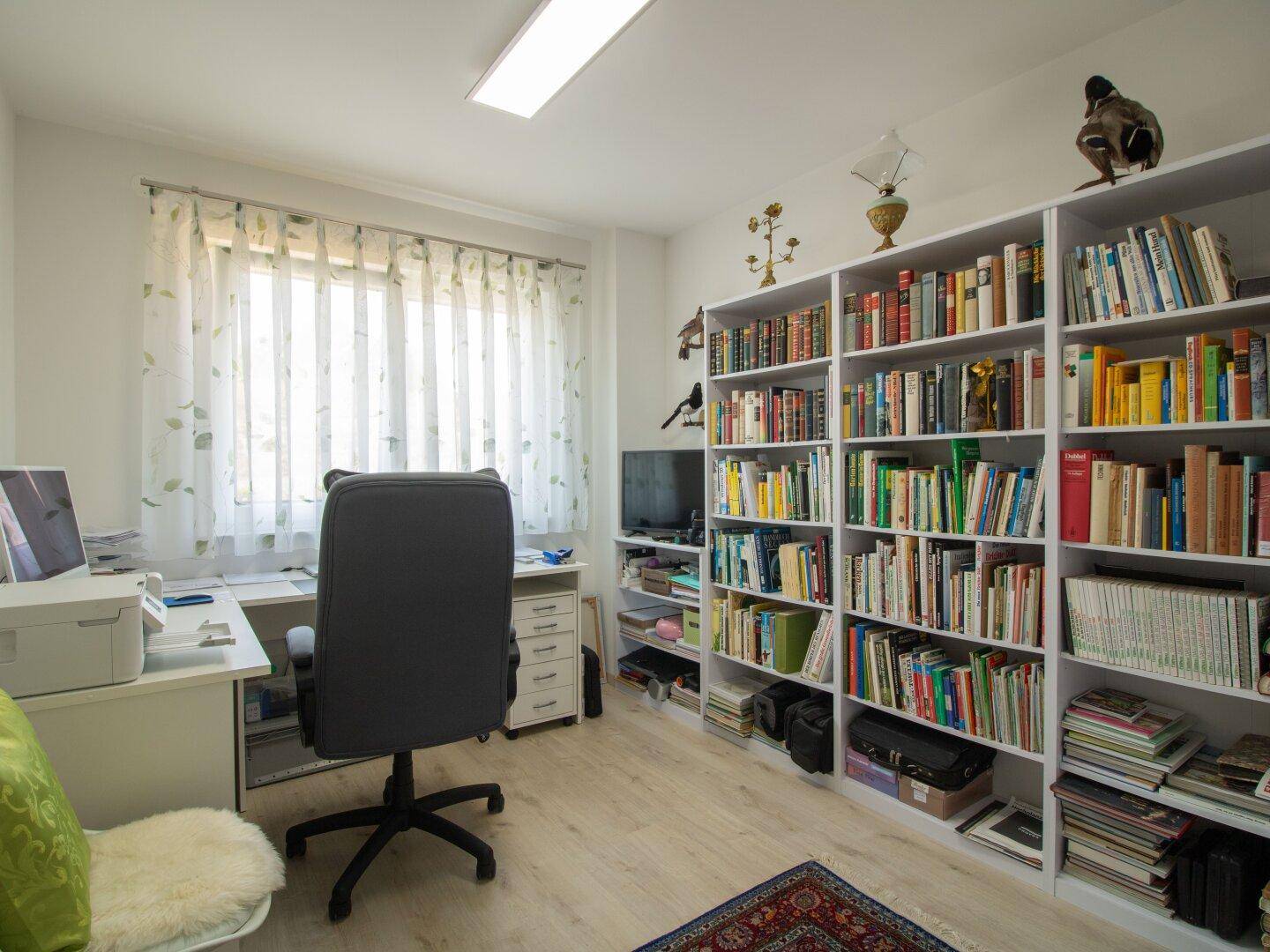 Arbeitszimmer mit großem Bücherregal, Schreibtisch und Fenster für natürliches Licht.
