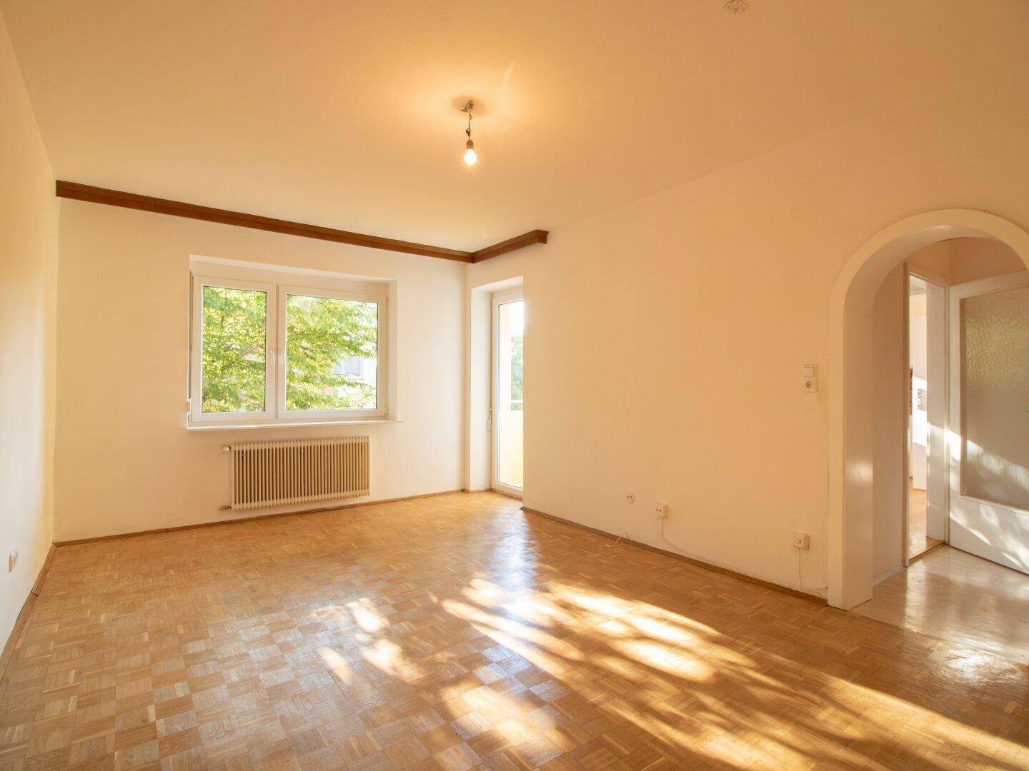 Helles, leeres Wohnzimmer mit Parkettboden, großem Fenster und Zugang zum Balkon.