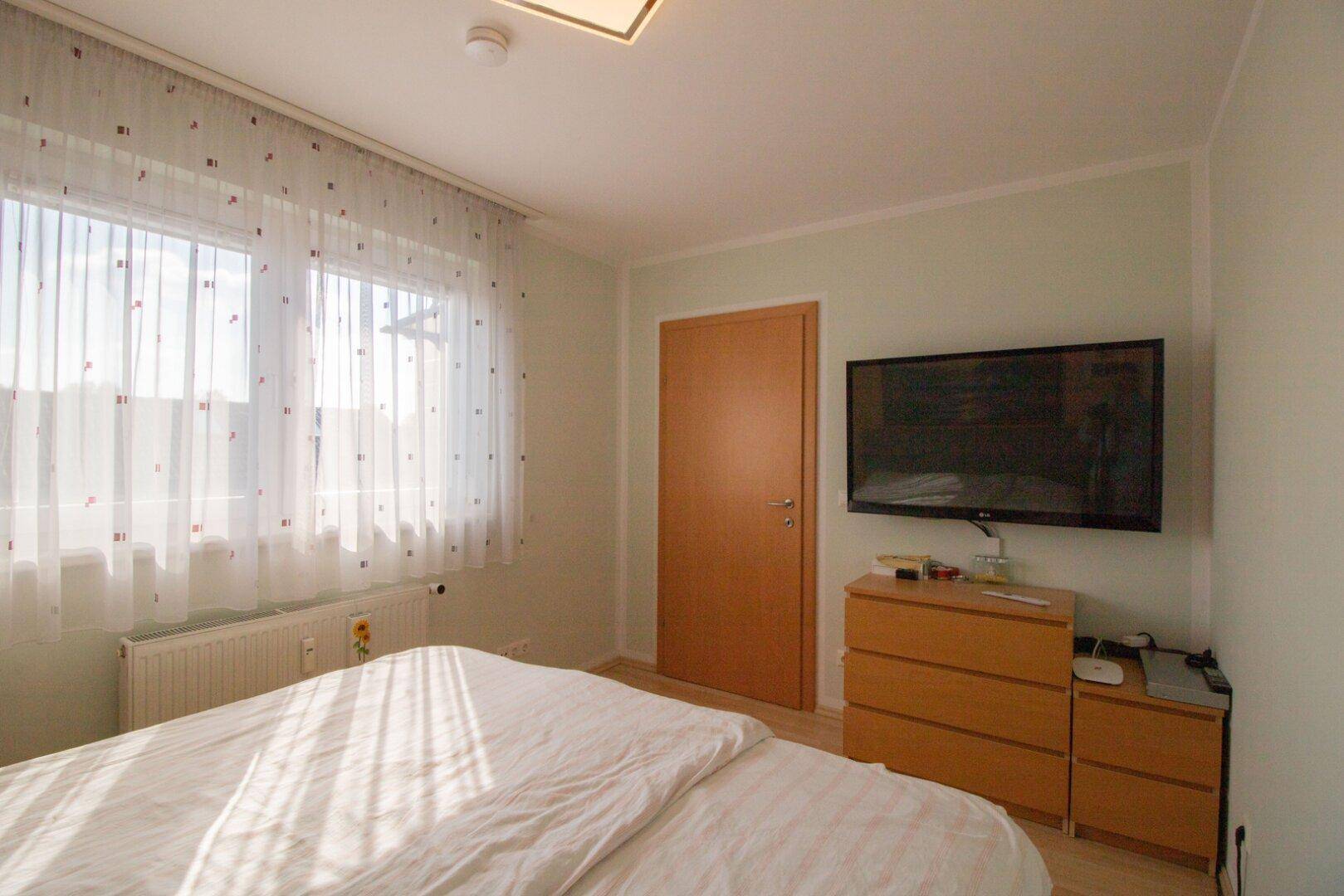 Helles Schlafzimmer mit Bett, Wandfernseher und Fenster, das für eine angenehme Atmosphäre sorgt.