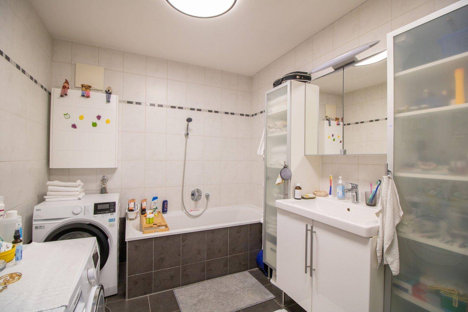 Helles Badezimmer mit Badewanne, Waschbecken und Waschmaschine, ausgestattet mit modernen Fliesen.