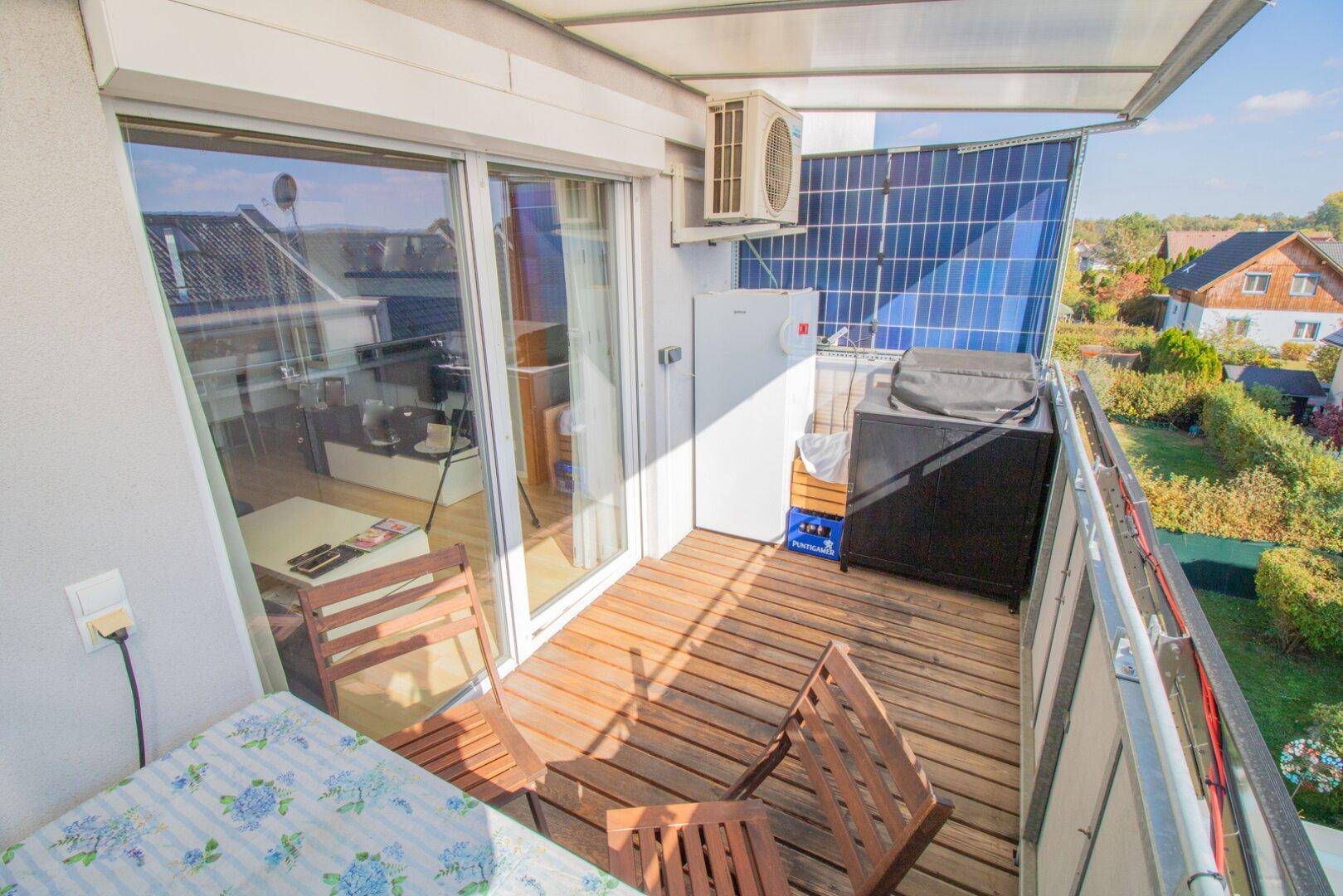 Sonniger Balkon mit Grill, Kühlschrank, Sitzgelegenheiten und Solarpaneelen mit Blick ins Grüne.