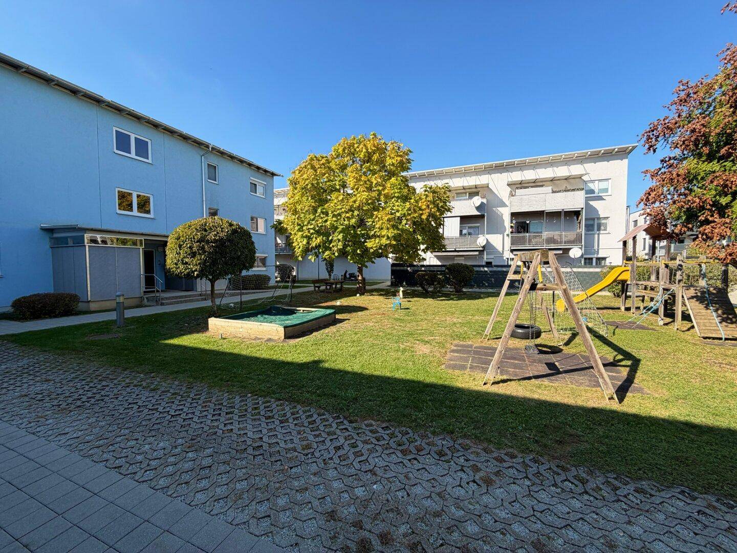 Grüner Gemeinschaftsgarten mit Spielplatz zwischen modernen Wohngebäuden unter blauem Himmel.