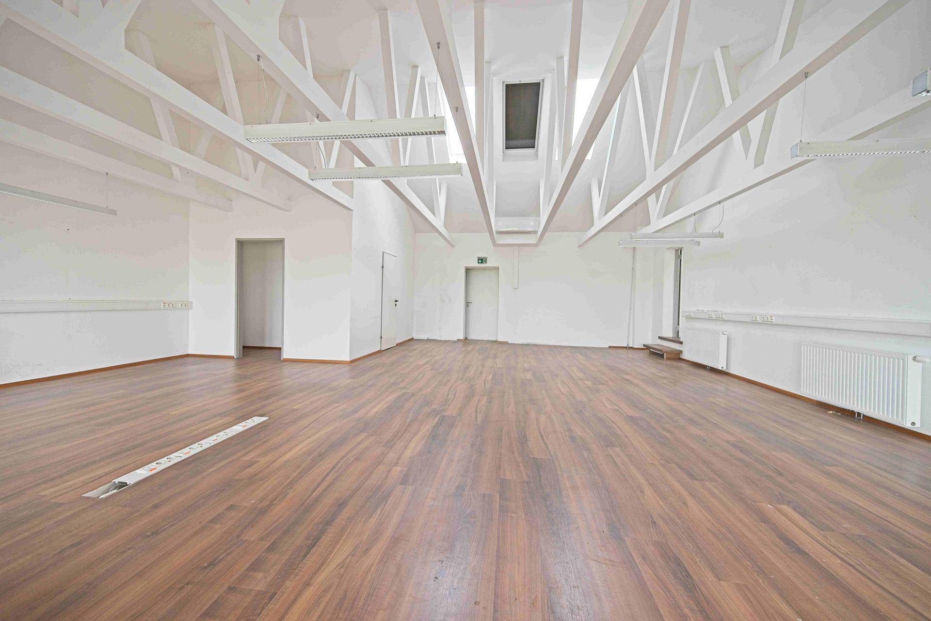 Großer, leerer Büroraum mit Laminatboden, weißen Wänden und einer beeindruckenden Deckenkonstruktion mit sichtbaren Balken.