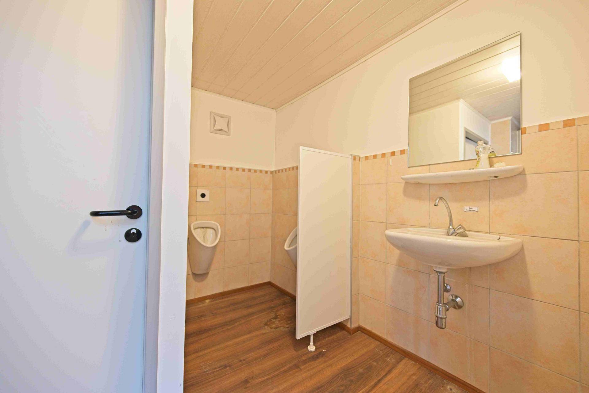 Herren-WC mit Urinalen, Waschbecken und beigefarbenen Fliesen an den Wänden.