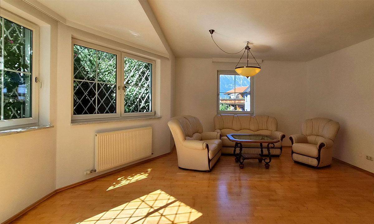 Helles Wohnzimmer mit Holzparkettboden, beigen Polstermöbeln und Fenstern mit Blick ins Grüne.