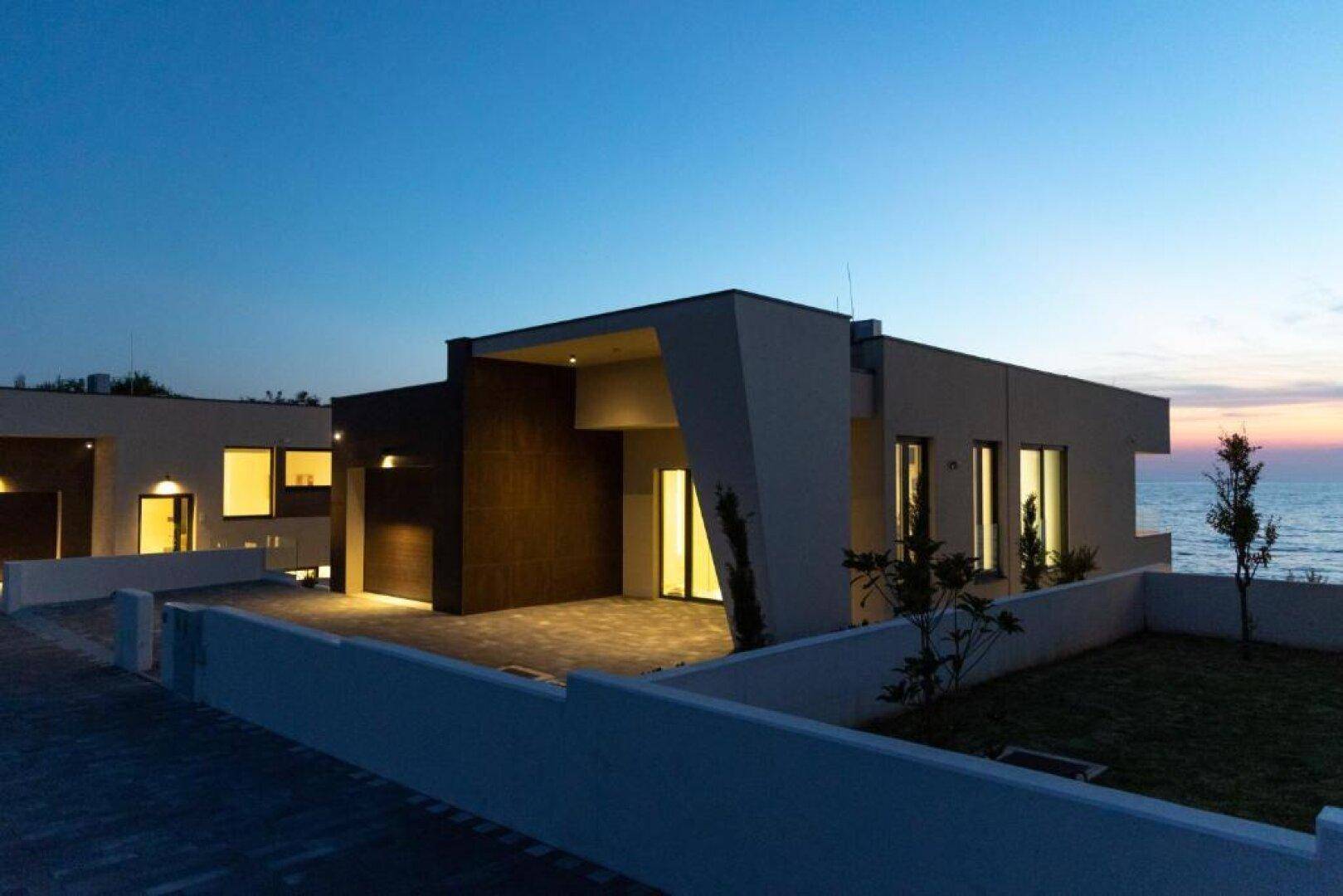 Moderne Architektur einer Villa bei Dämmerung mit Meerblick und beleuchtetem Eingangsbereich.