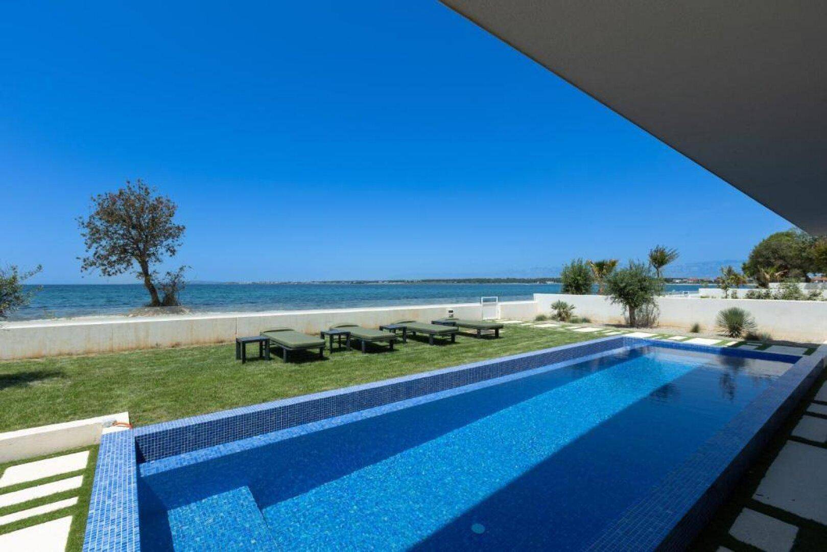 Langer, blauer Pool mit Mosaikfliesen und direktem Blick auf das Meer und den Strand.