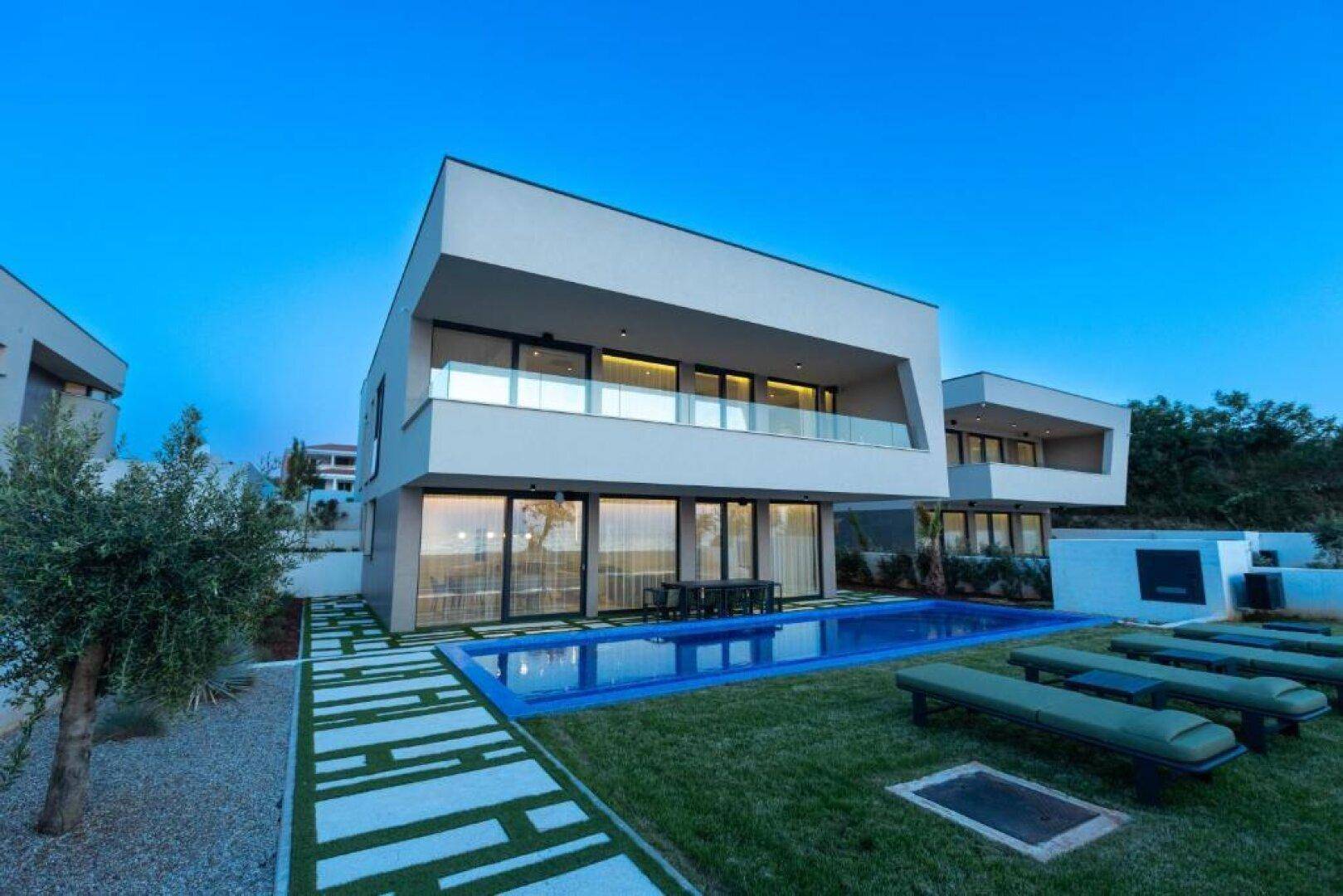 Elegante Villa bei Sonnenuntergang mit beleuchtetem Pool und Liegestühlen im Garten.