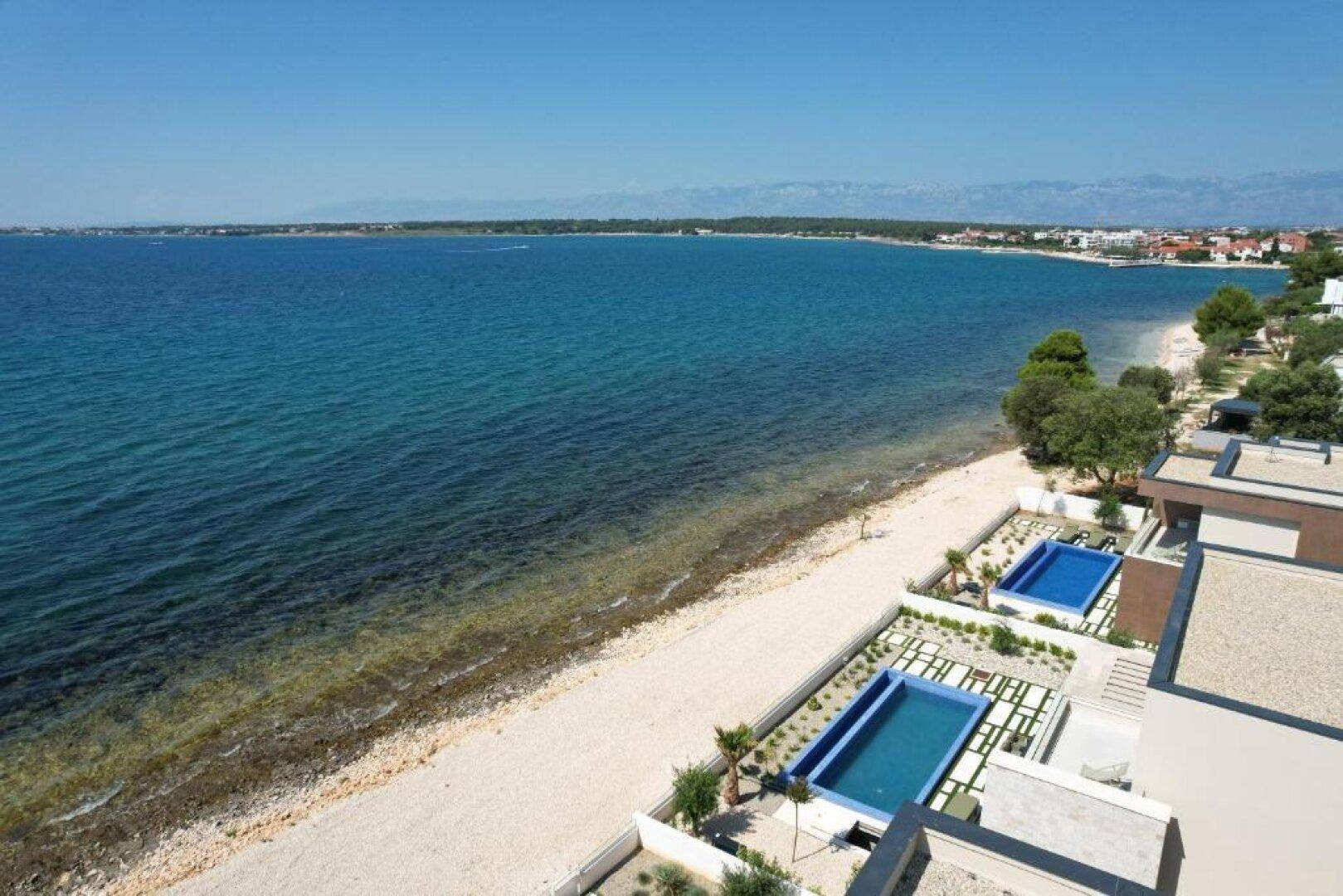 Luftaufnahme der Küstenlinie mit modernen Villen, Pools und dem weiten, blauen Meer.