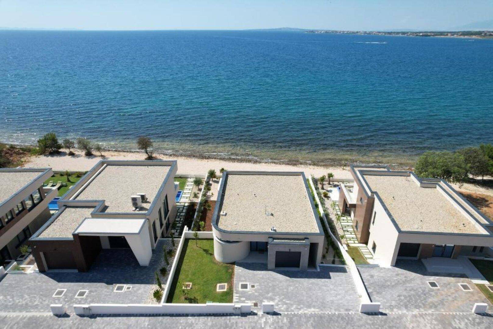 Luftaufnahme von mehreren modernen Villen direkt am Meer mit flachen Dächern und Pools.