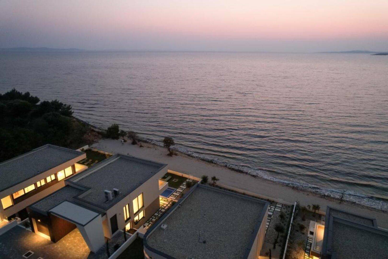 Abendliche Luftaufnahme der Küste mit Villen und dem ruhigen Meer bei Sonnenuntergang.