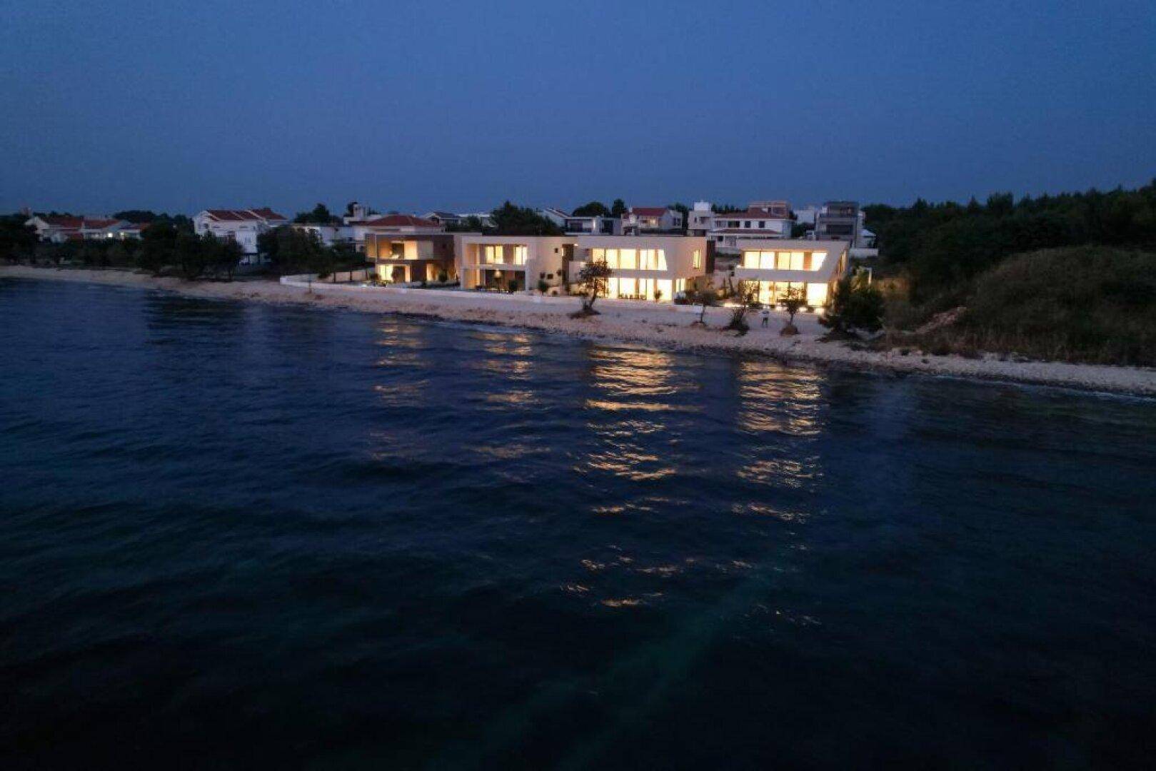 Nachtansicht der beleuchteten Villen vom Meer aus, mit reflektierenden Lichtern auf dem Wasser.