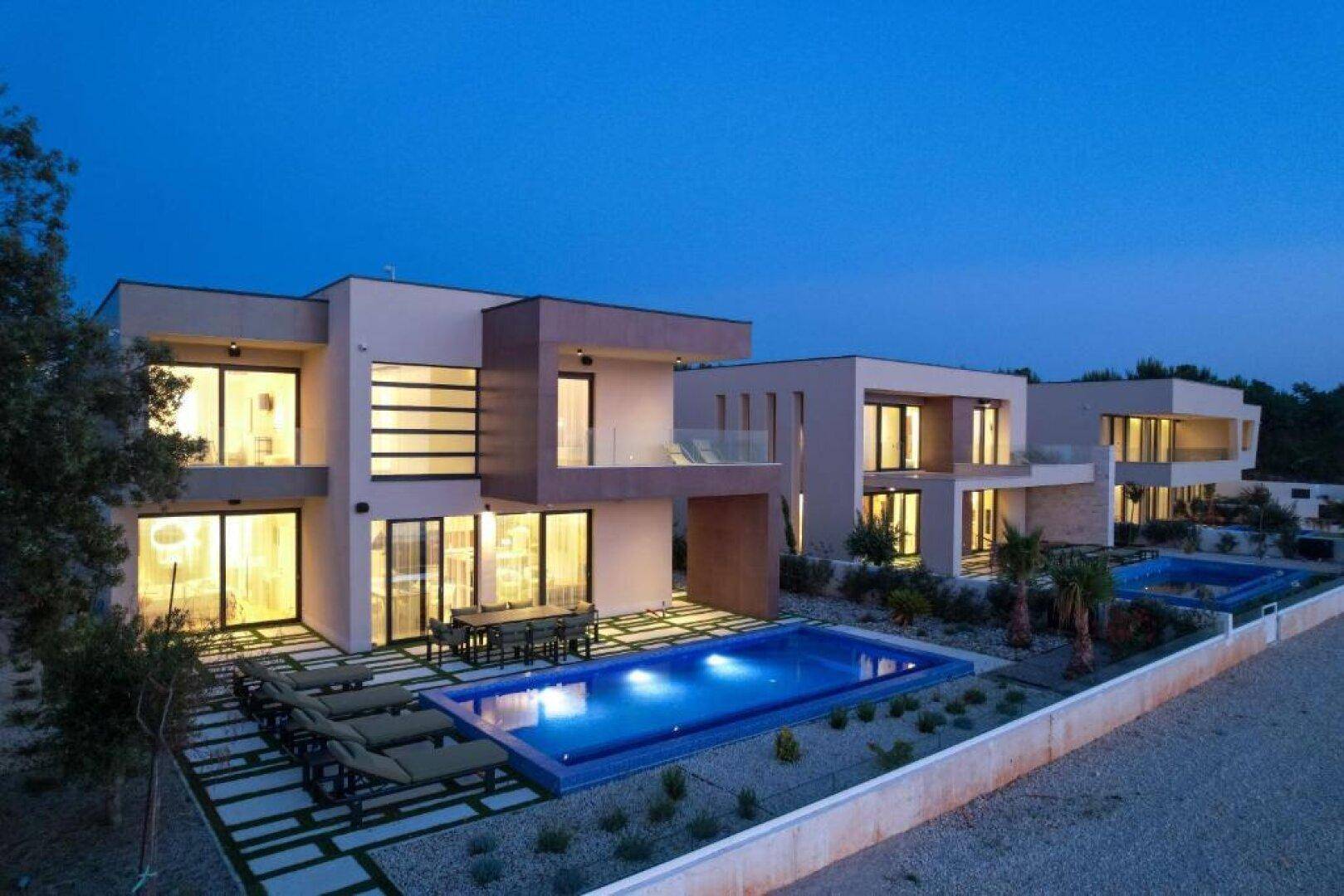 Reihe moderner Villen mit beleuchteten Pools und Terrassen, die eine luxuriöse Wohnanlage bilden.