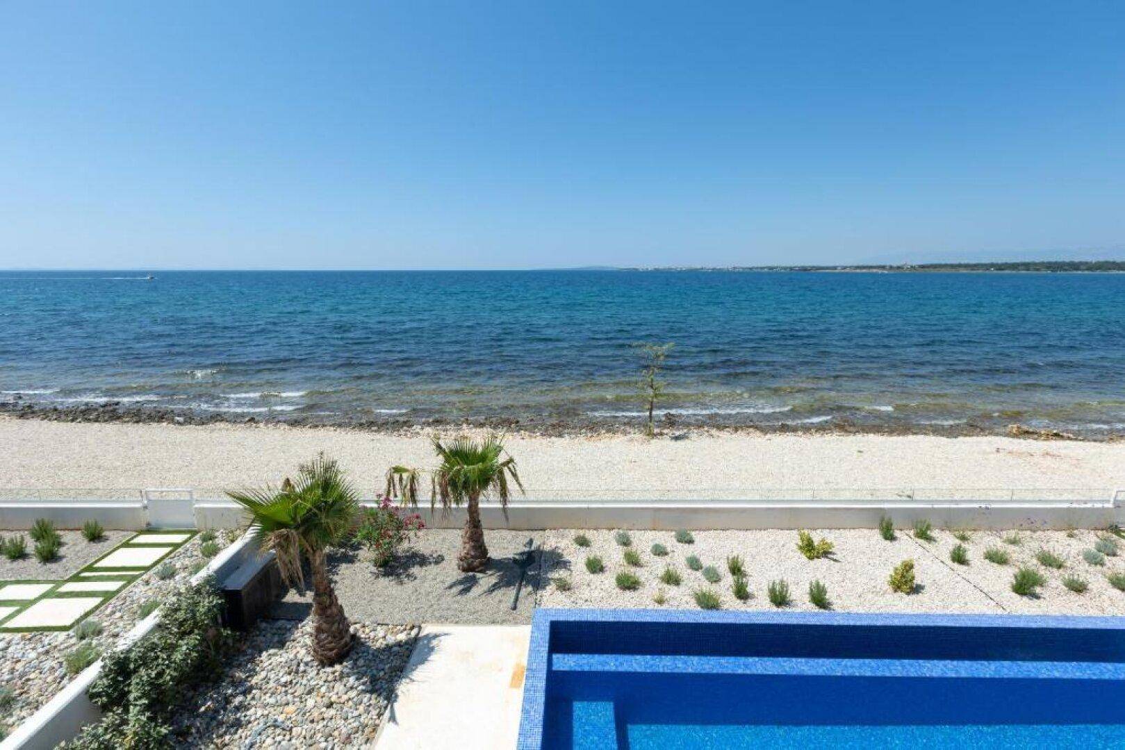 Direkter Blick auf das klare Meer und den Kiesstrand, umgeben von mediterraner Bepflanzung.