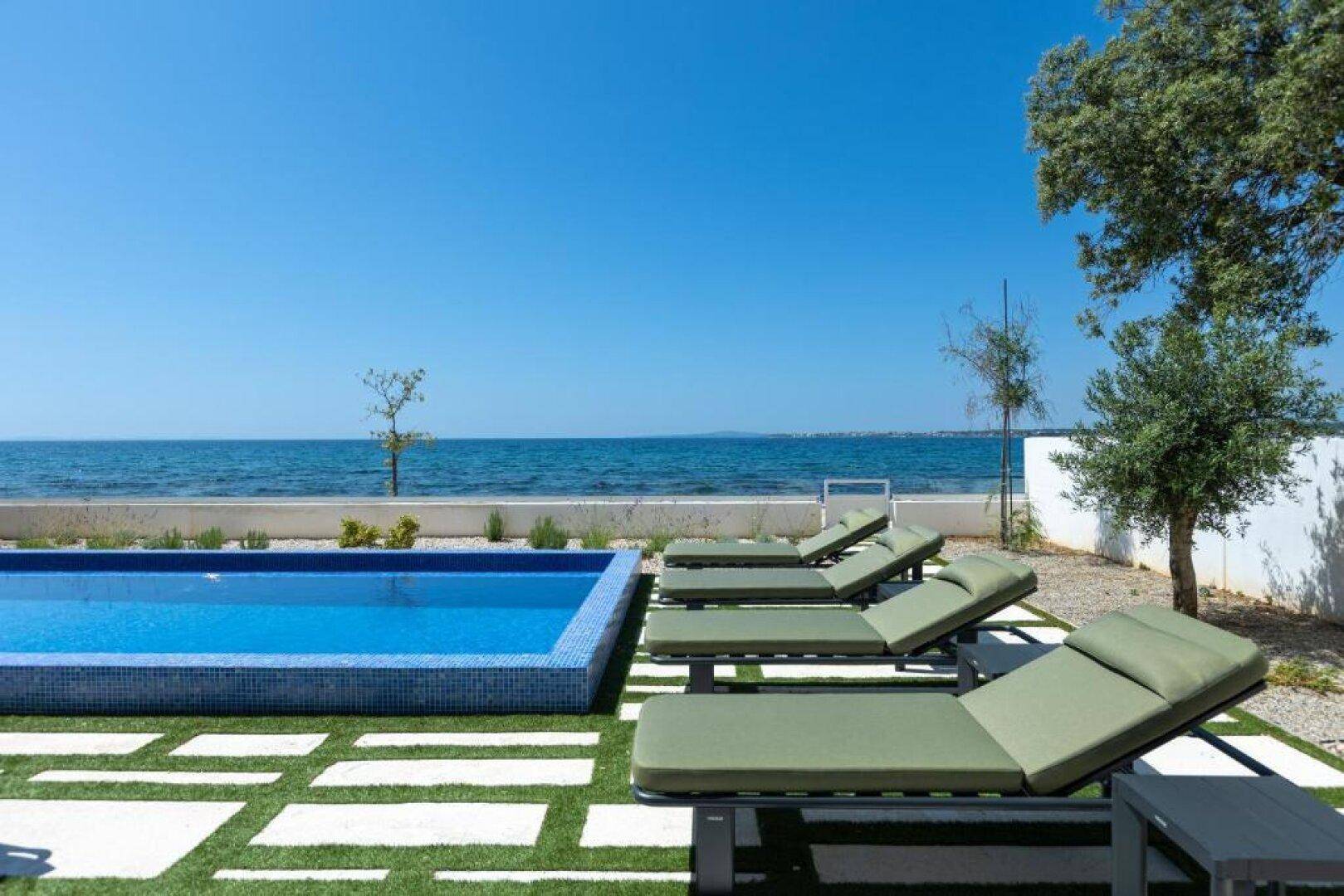 Entspannender Poolbereich mit Liegestühlen und direktem Zugang zum Meer, umgeben von Grünflächen.
