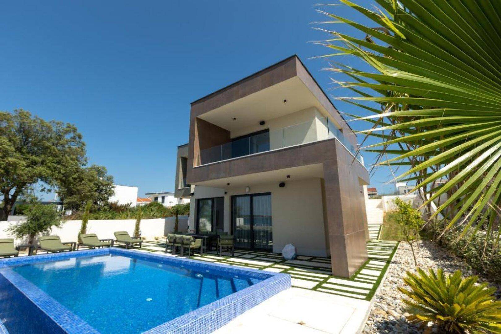 Moderne Villa mit Pool und Liegestühlen unter blauem Himmel, umgeben von mediterraner Vegetation.