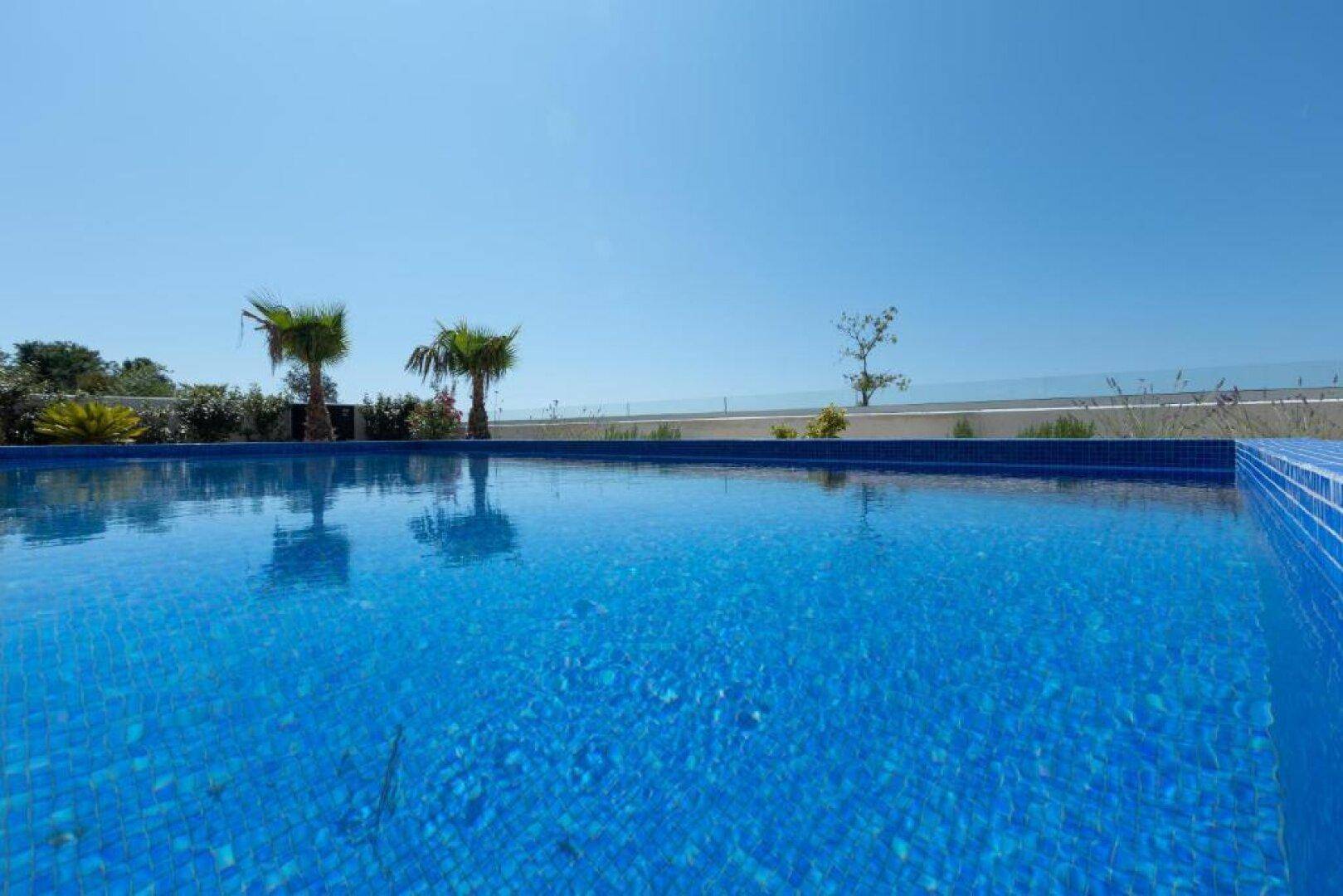 Nahaufnahme des blauen Pools mit Meerblick, umgeben von mediterraner Vegetation und Palmen.