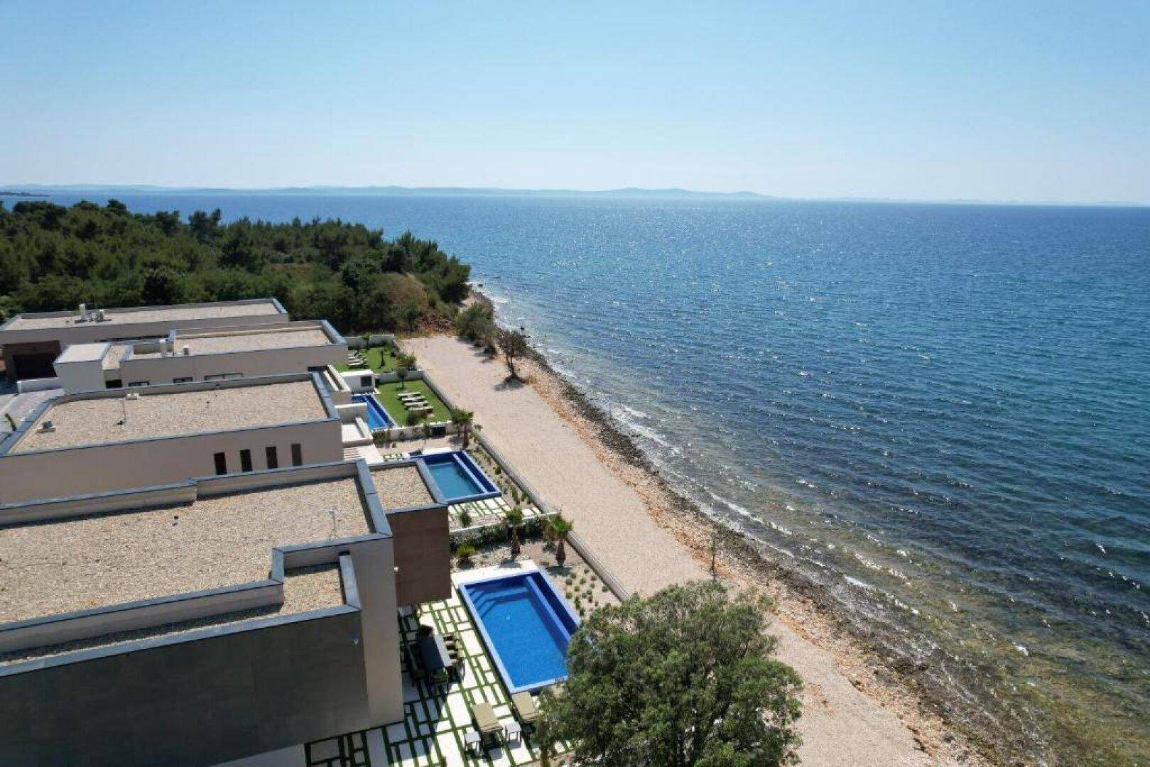 Luftaufnahme moderner Villen mit Pools und Gärten, direkt am Strand gelegen, mit weitem Meerblick.