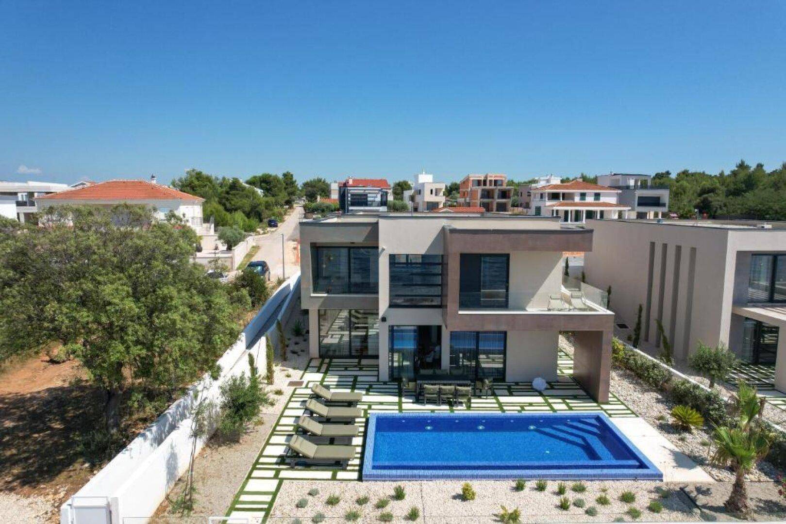Luftaufnahme der modernen Villa mit Pool und Garten, umgeben von weiteren Häusern und Grünflächen.