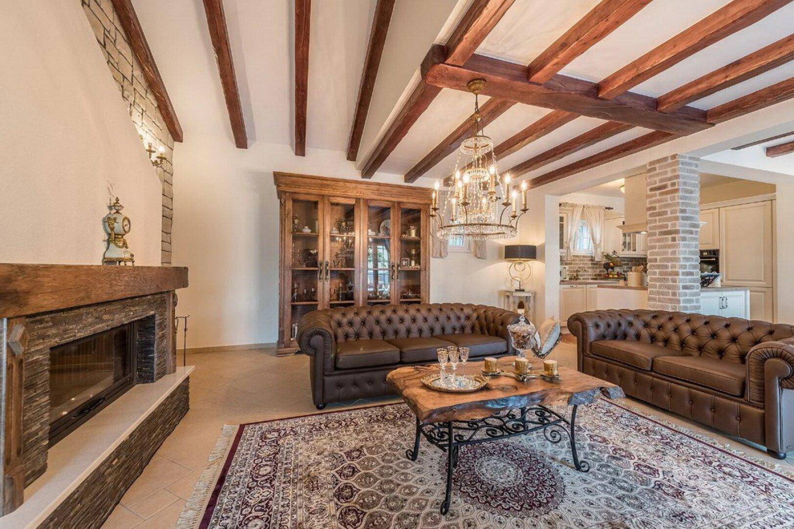 Geräumiges Wohnzimmer mit Kamin, Ledersofas und einem großen Teppich, unter einer Holzbalkendecke.