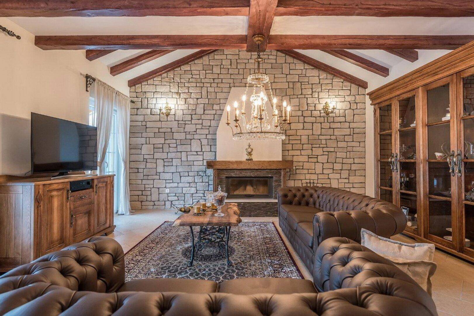 Einladendes Wohnzimmer mit Steinkamin, Ledersofas und einem eleganten Kronleuchter unter der Holzbalkendecke.
