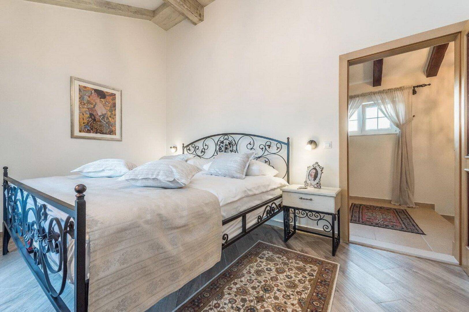 Schlafzimmer mit schmiedeeisernem Doppelbett und Blick in den angrenzenden Raum, unter einer Holzbalkendecke.