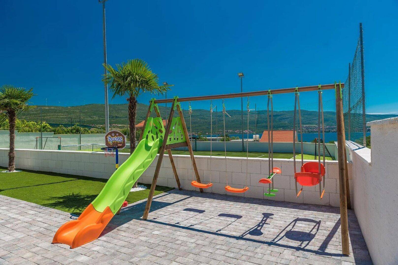 Kinderspielplatz mit Rutsche und Schaukeln auf Kunstrasen, umgeben von einer niedrigen Mauer.