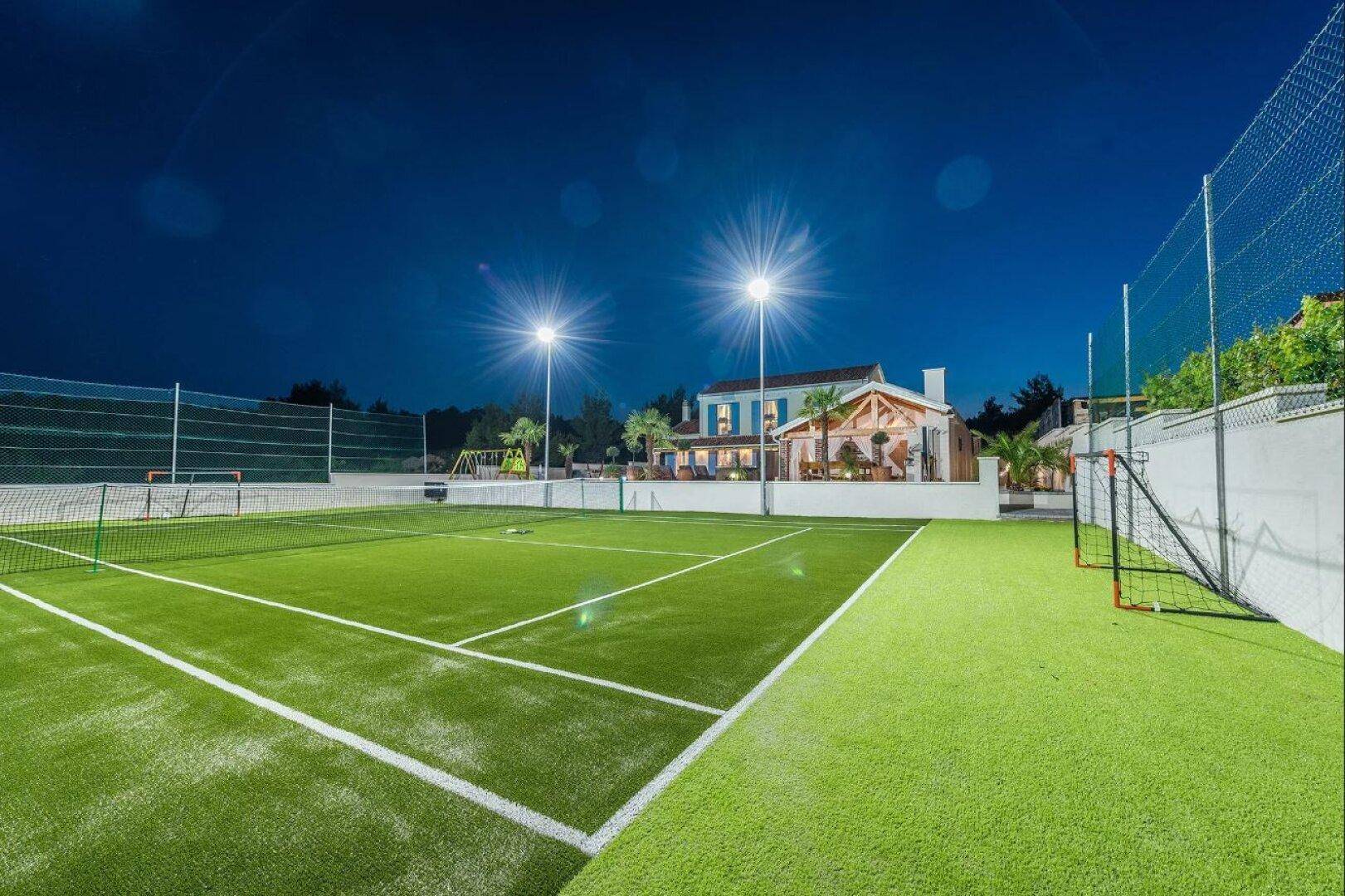 Beleuchteter Tennisplatz mit Kunstrasen und Fußballtoren, mit der Villa im Hintergrund bei Nacht.