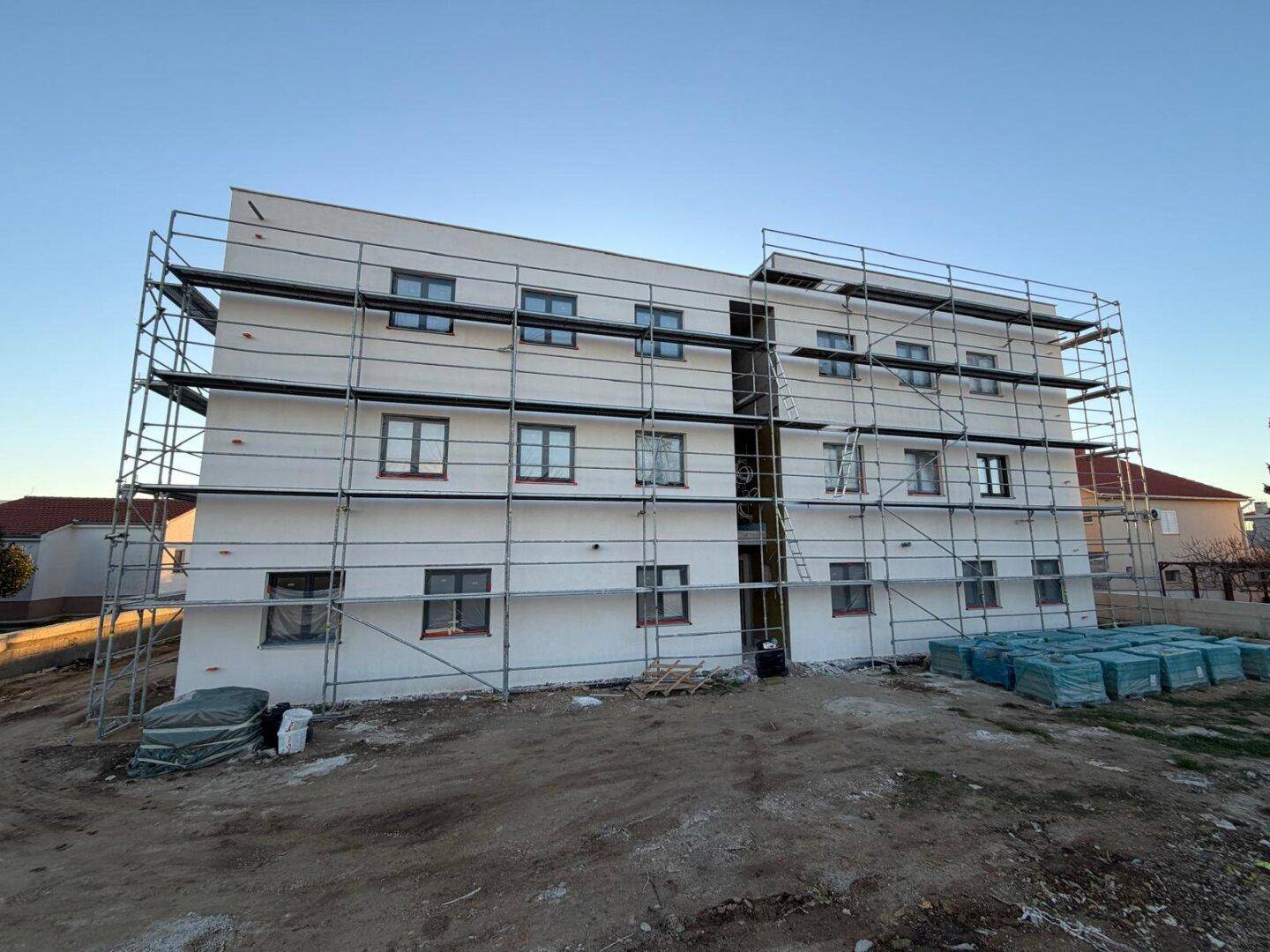 Fassade eines im Bau befindlichen Gebäudes mit Gerüst und Baumaterialien im Vordergrund der Baustelle.