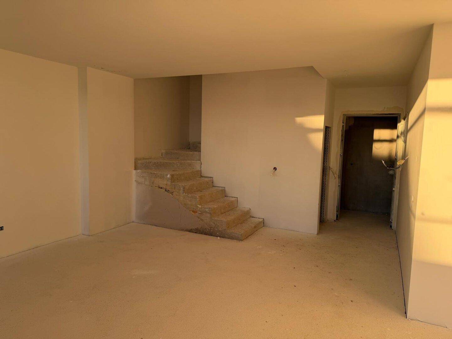 Rohbau eines Innenraums mit Betonwänden und einer unfertigen Treppe, die auf die nächste Etage führt.