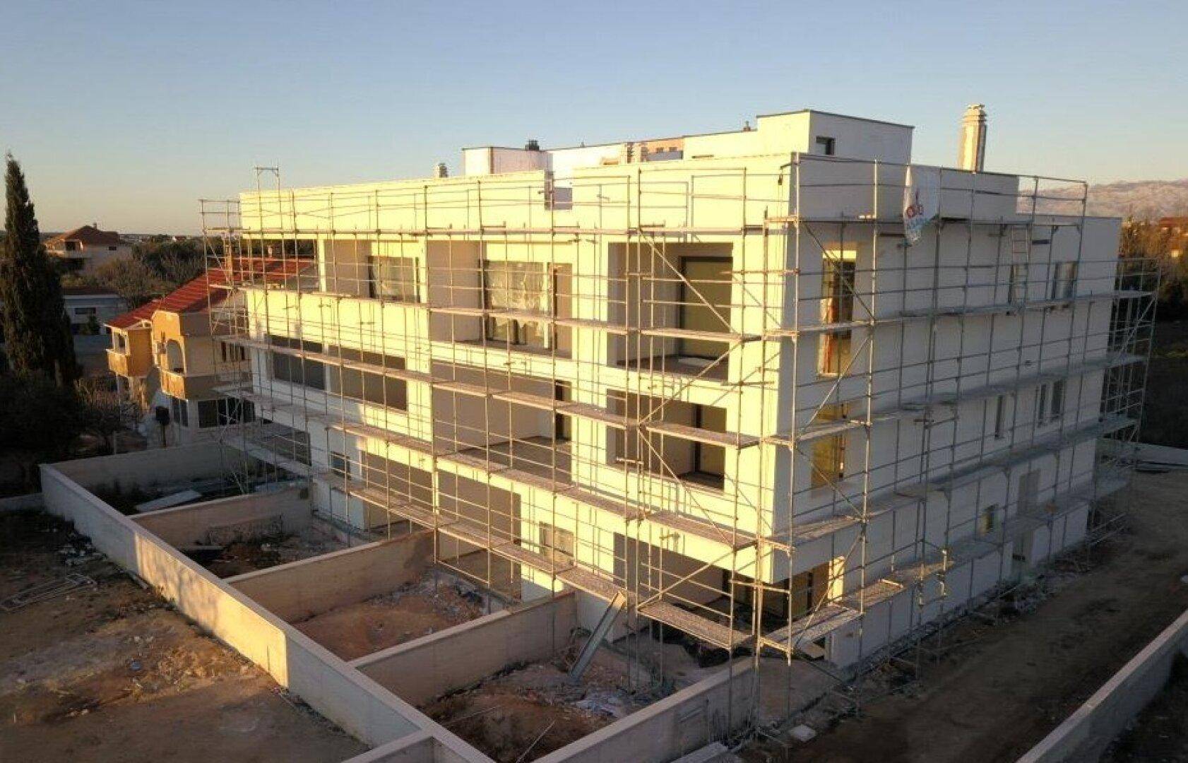 Gebäude im Bau mit Gerüst, das die Fassade bedeckt, und sichtbaren Fundamenten im Vordergrund.