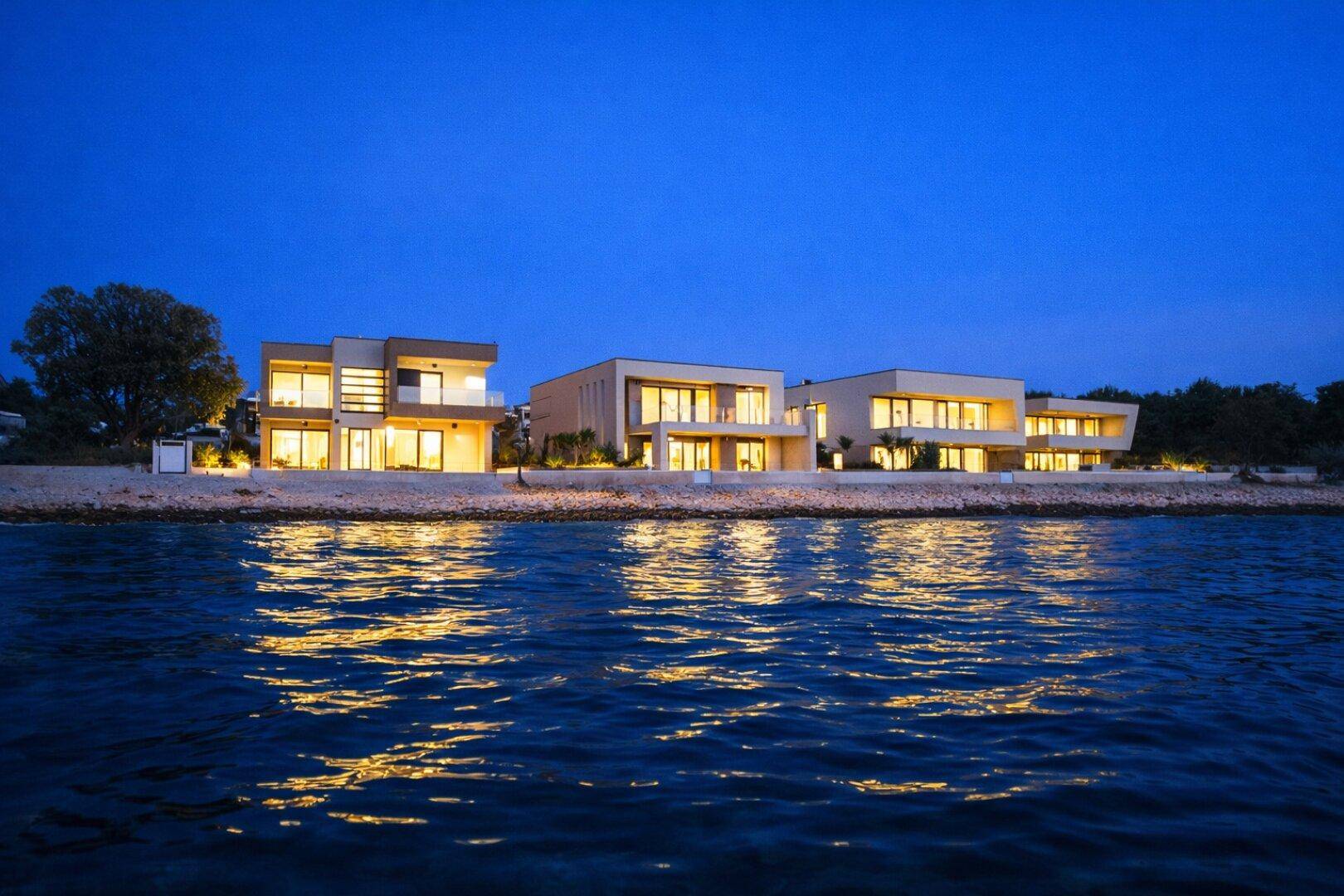 Reihe moderner Villen direkt am Meer, beleuchtet in der Abenddämmerung mit Reflexionen im Wasser.