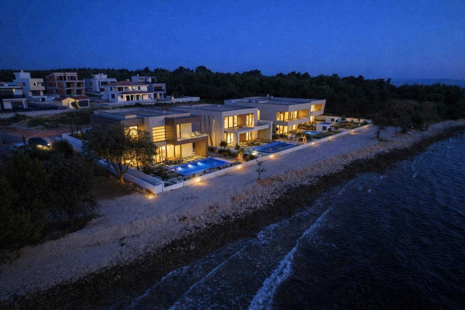 Luftaufnahme eines modernen Villenkomplexes mit Pools und Meerzugang bei Nacht.
