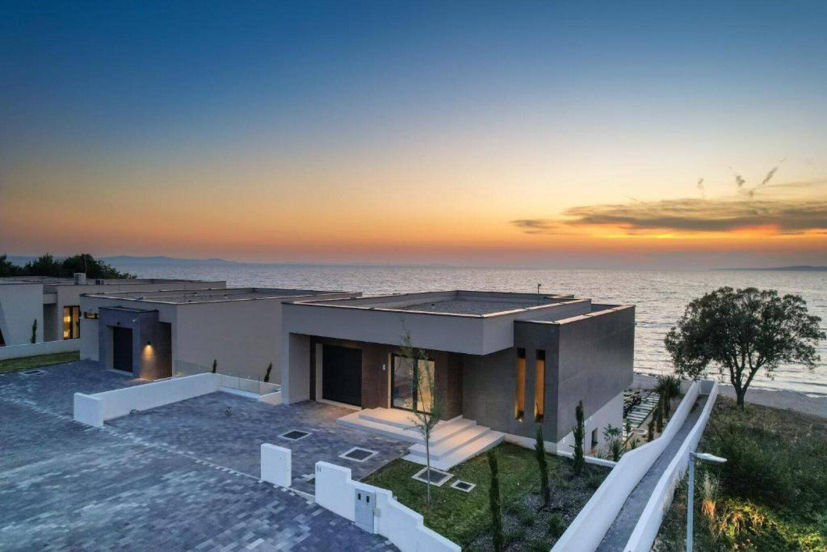 Moderne Villa mit Flachdach, großem Vorplatz und Meerblick bei Sonnenuntergang.