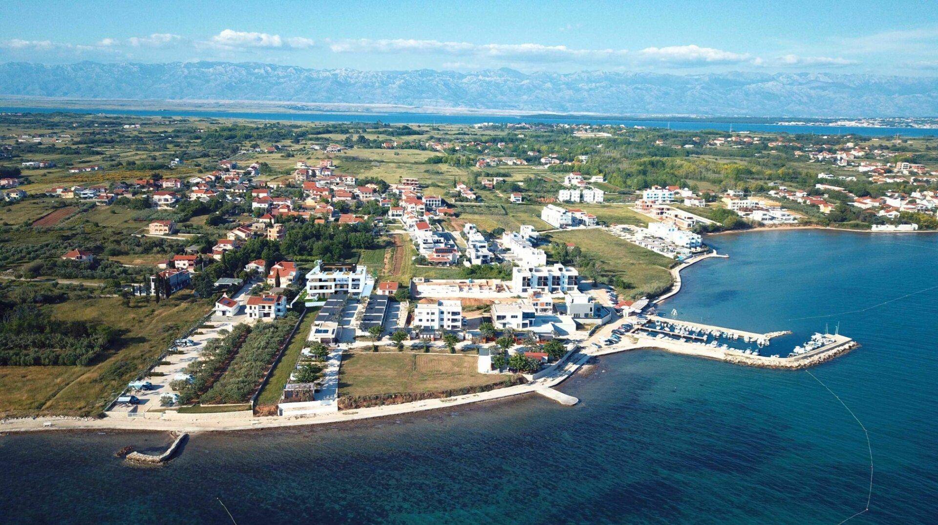 Luftaufnahme einer Küstensiedlung mit Yachthafen, umgeben von Meer und grüner Landschaft.